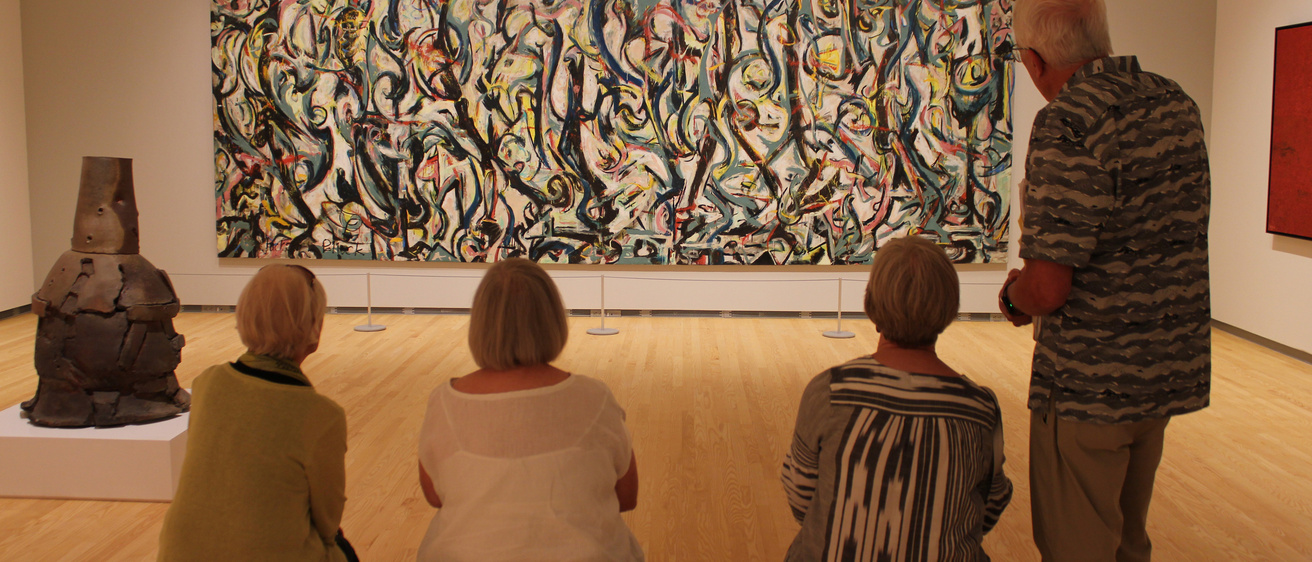 Four people observe an abstract painting in a gallery. One person stands while three sit on a bench. Nearby is a metal sculpture on display.