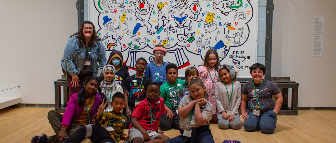 A group of children smile for a photo in front of a colorful mural painting in a gallery