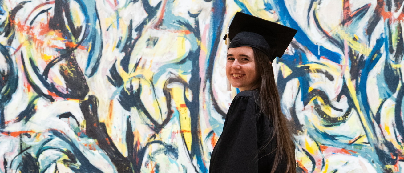 Gallery Host and Campus Engagement Coordinator Annelies Knight poses in her graduation gown in front of Jackson Pollock's Mural.