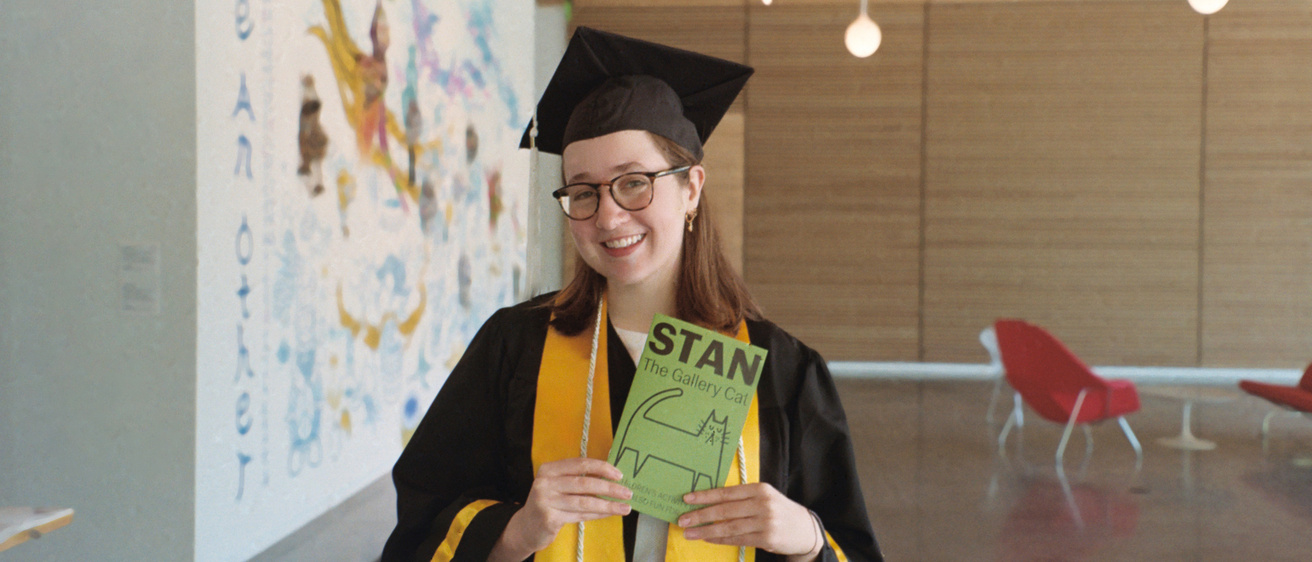 Museum Programs Intern Abbie McLaren, dressed in her graduation attire, poses with a Stan the Cat activity booklet, which she created in the spring of 2025, in the Stanley Museum of Art lobby.