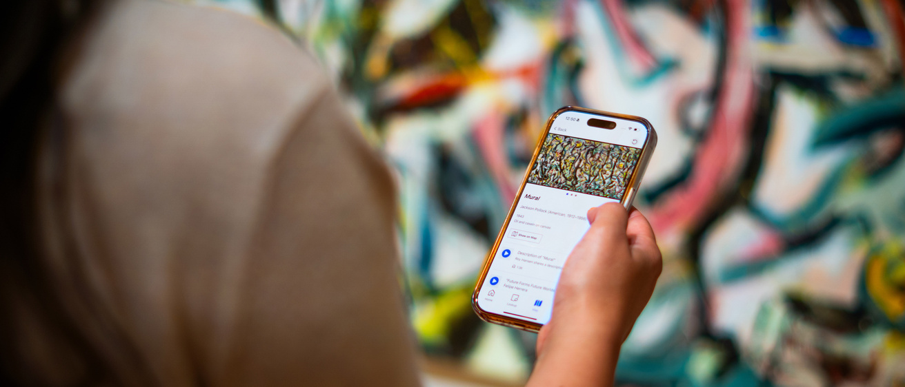A close up of persons shoulder and hand. They are holding up a phone with a colorful abstract painting on the display screen and some text. In the background is the same painting as the one on the phone.