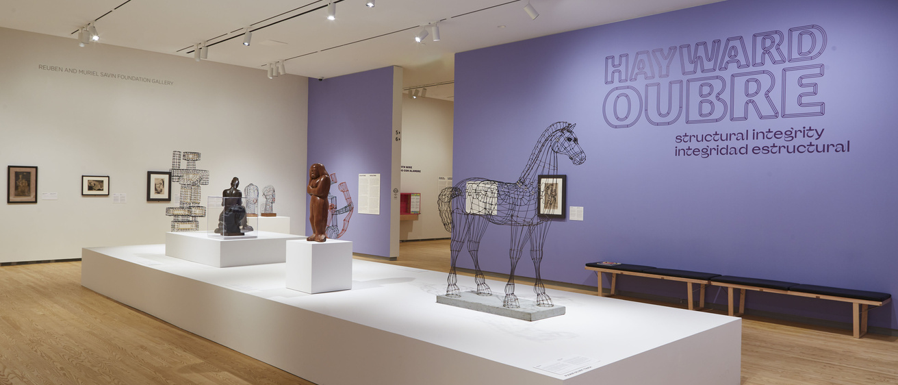 An open gallery space with light wooden floors displays wire and metal sculptures on white platform in the center of the room, with purple accent walls and the text "Hayward Oubre: Structural Integrity" featured prominently on the wall.