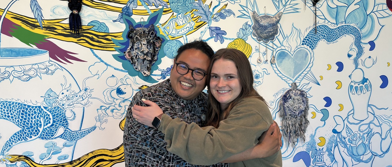 Shaun and Amelia hug each other, posing in front of the Jiha Moon mural in the museum lobby
