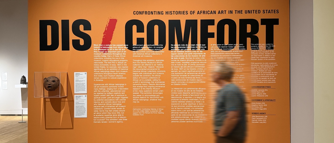 Entrance wall for the Stanley Museum's Dis/Comfort exhibition, with the title in large black letters split by a red slash, bilingual introductory text on an orange background, a Makonde mask displayed in a wall case, and a visitor walking past.