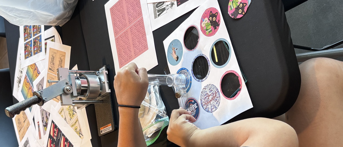 A student sits at a table, cutting out circular pieces of paper to use with the button maker.