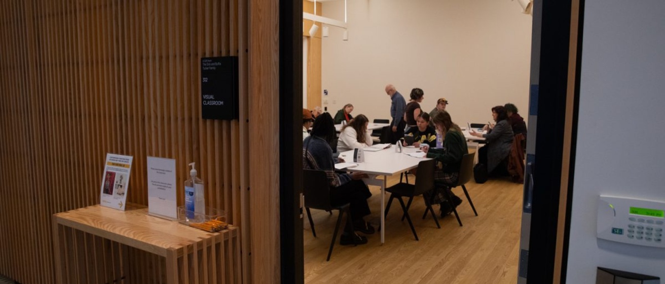 A group of people sit around white tables in a bright classroom, talking and working on paper, seen through an open doorway.