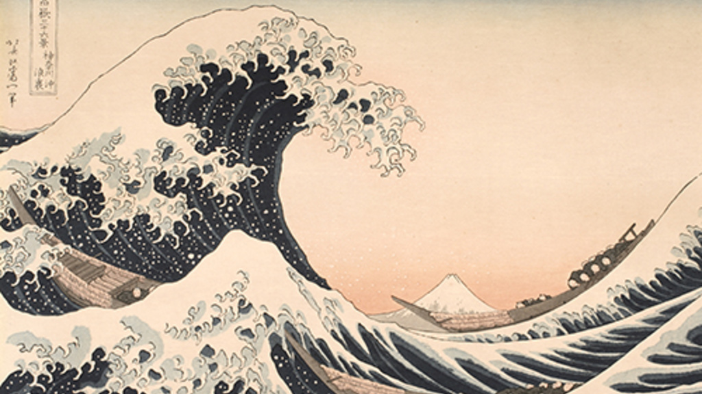 Woodblock print of a large dark blue wave with frothing white caps. The crest rises from the left and reaches out over a mountain in the middle which appears small and distant. Three long boats with people in grey robes ride the wave.
