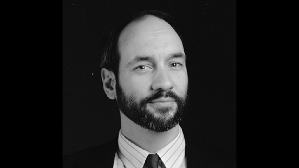 Black and white portrait of man with beard and moustache wearing dark blazer, white shirt, and dark tie