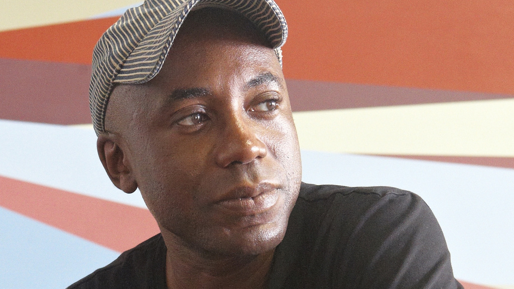 A Black man in a dark t-shirt and beige ball cap in front of a mural painted with geometric shapes in bright colors.