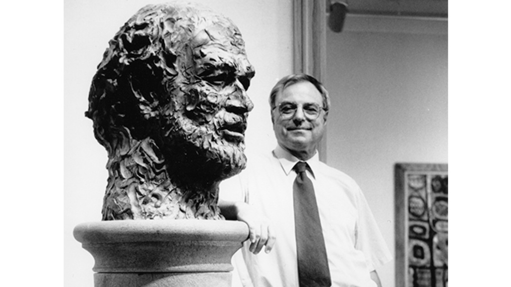 Man with glasses wearing a white shirt and dark tie stands looking at viewer with his elbow resting on a pedestal supporting a sculpture of a bearded man's head