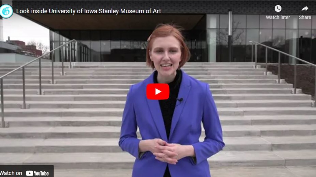 Woman with short red hair wearing a bright blue jacket stands at the foot of a staircase leading up to the Stanley Museum of Art