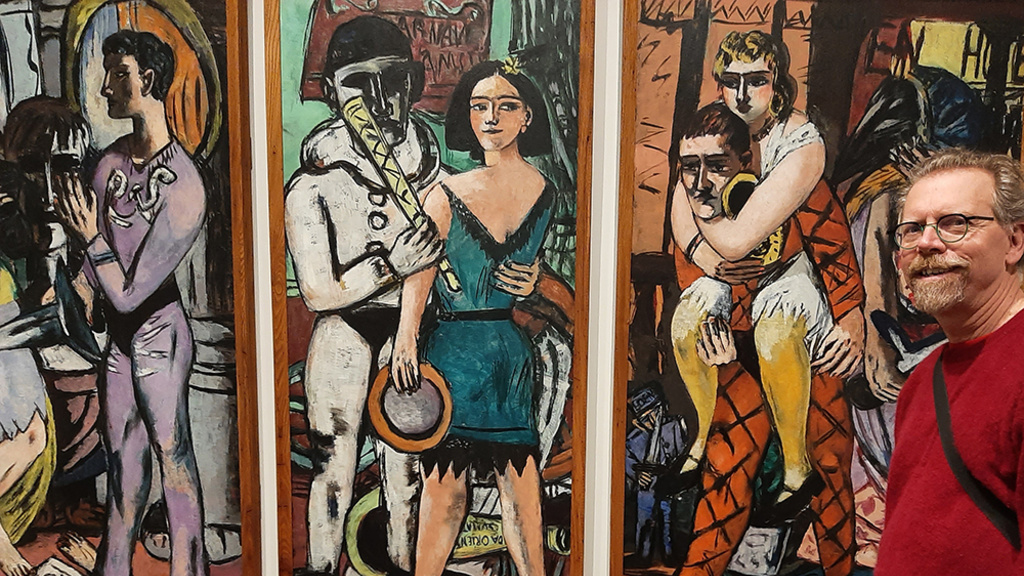 A man with beard and glasses stands in front of a three-panel painting. L: Person in yellow dress stands, one leg crosses the other, plays a flute next to figure in purple holding a knife. Behind is large sculpted head, partly in shadow. Cntr: Person in black and blue dress clutches circular object, holds long cylindrical object like a horn, stands close to figure in a black and white clown costume.  R: Tired harlequin clown carries a jewelry clad person piggyback.