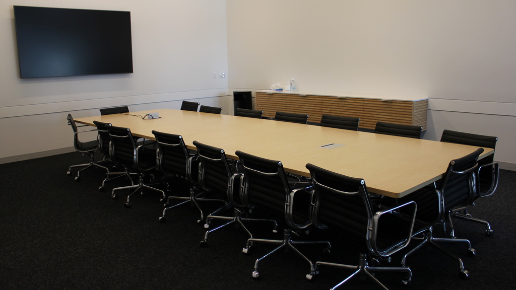 A meeting room with a long, wooden table surrounded by 16 black chairs. There is a large monitor mounted to the wall at left and a credenza along the far wall.