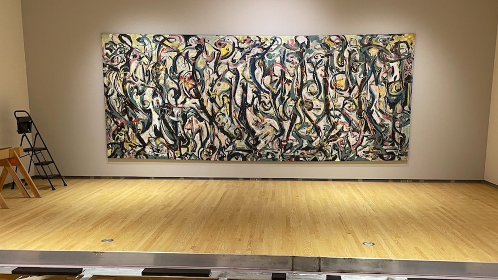 A large abstract painting with swirls of yellow, pink, teal, and light greenish blue, amid longer, vertical, curved black lines that have a quality of dance-like movement shown on a gallery wall with an aluminum travel frame on the floor in foreground.