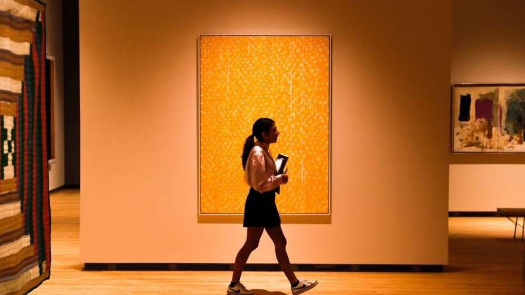 A woman with a short skirt and ponytail walks in front of a large painting on the wall of a museum gallery