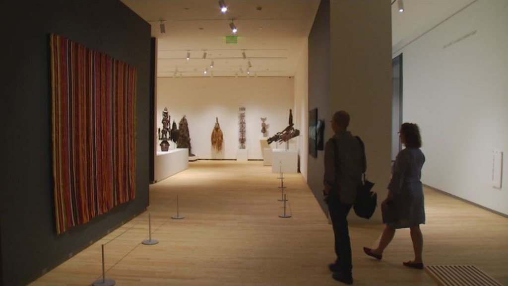 Two people walk through a museum gallery with textiles on the left and African masks in the background