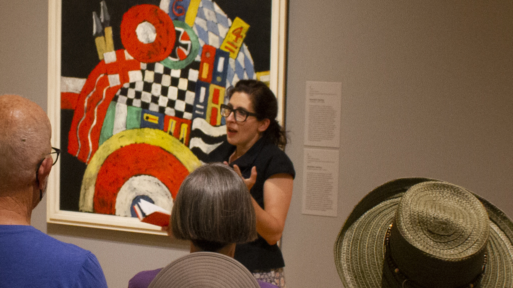 A female art museum curator stands in front of a brightly colored, geometric painting speaking to a group of visitors