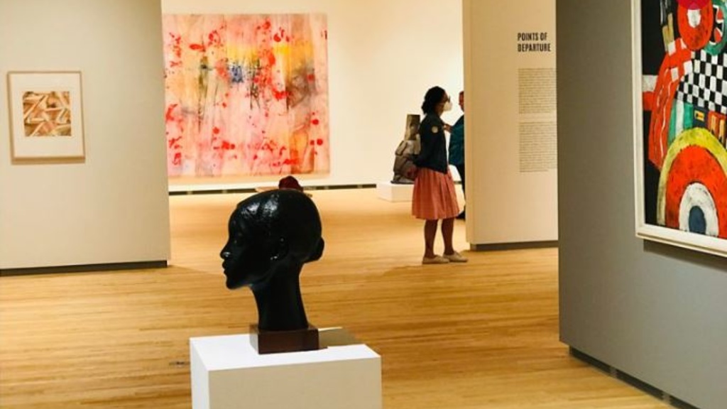 An art gallery with a bronze bust in the foreground, a brightly colored, geometric painting on the right, and a person viewing a large multi-colored abstract painting in the background