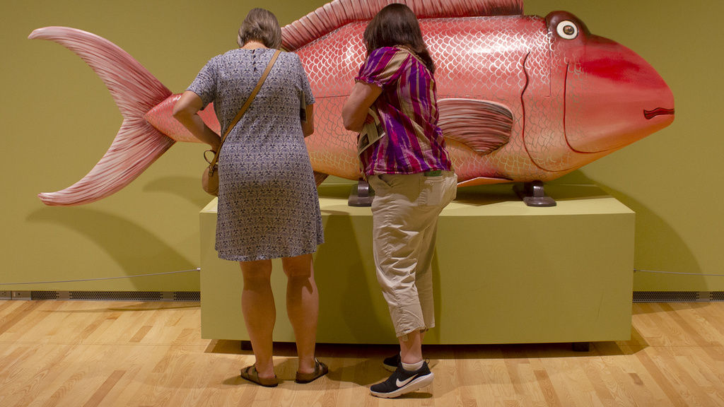 Two women, seen from behind in a green room with wooden flooring, look at a large, pink, wooden fish sculpture.