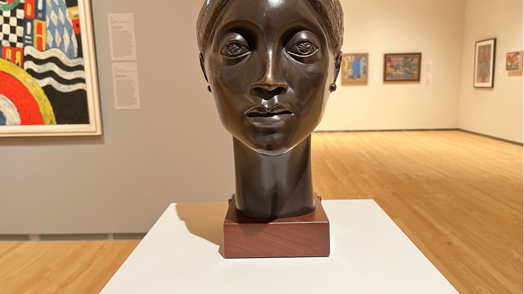 A bronze bust of a female with hair pulled back tightly rests on a white pedestal in an art gallery