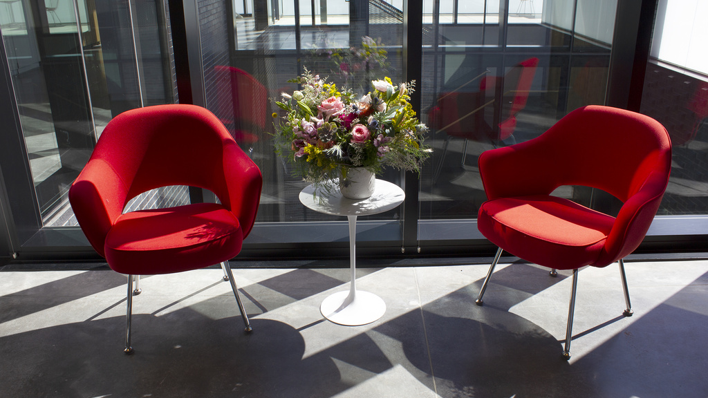 Two red armchairs and a small table with flowers sit in front of a glass window with sunlight streaming in.