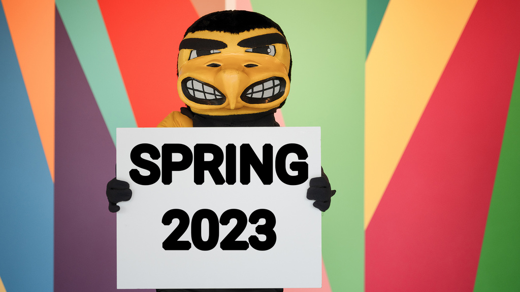 Herky the Hawk mascot holding a sign "Spring 2023" while standing against a brightly colored wall mural