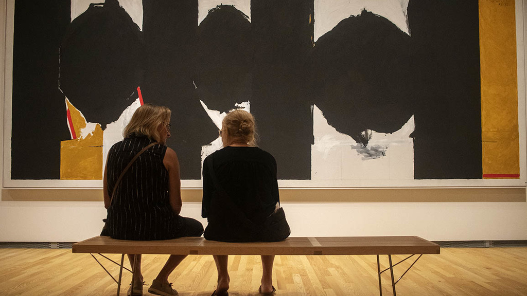 Two women sit on a wooden bench in front of a large wall painting