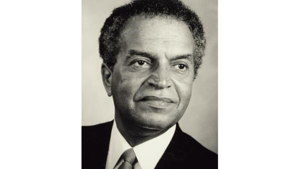 A black and white portrait of a Black man wearing a dark jacket with a white shirt and tie.