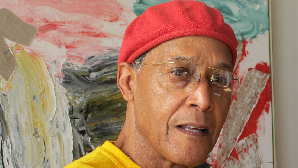A Black man wearing a red beret, bright yellow sweatshirt, and wire-rimmed glasses stands in front of an abstract painting