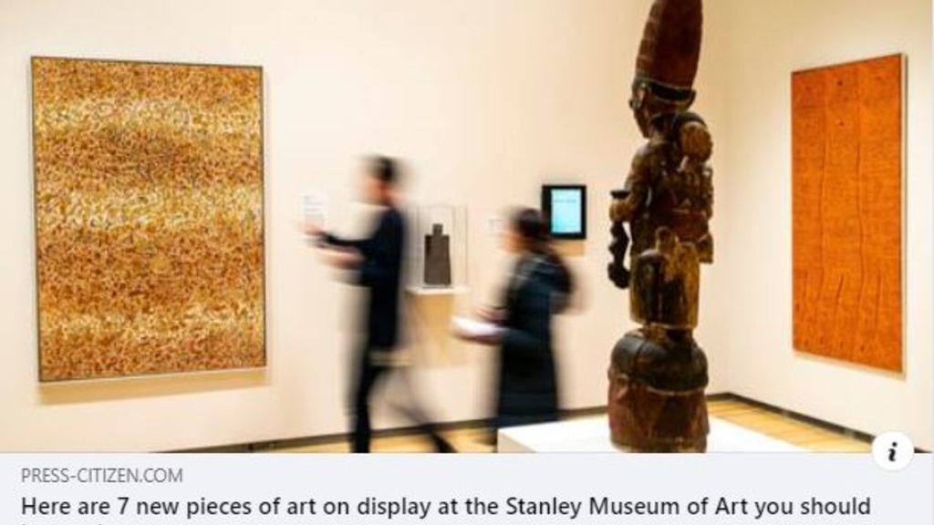 An art gallery with a tall, wooden sculpture at center and two abstract, earth-toned paintings at rear. There are blurred figures walking past the art.