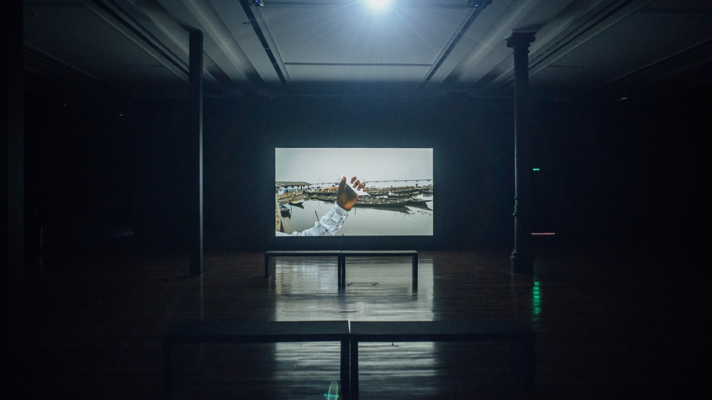 An installation image of a video work; the room is dark, with benches and pillars visible in shadow. At the center of the image is a screen, displaying a still from a video in which an arm is outstretched, hand holding a cell phone with the reflective surface lit with light, in front of a background of a shipyard, with large boats and water visible.