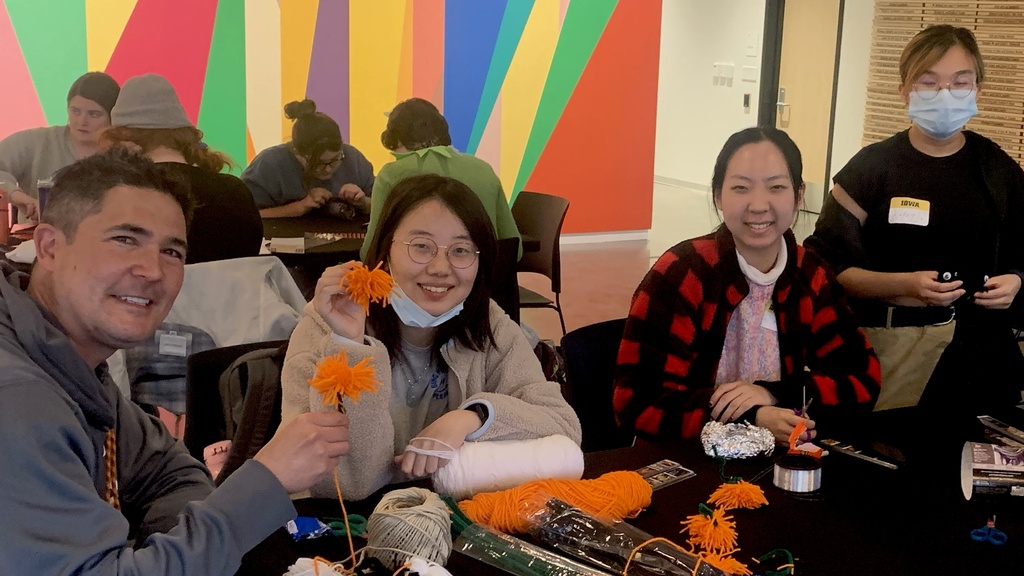 A photo of four students gathered around a folding table in the Stanley Museum of Art lobby. They are all in the midst of making crafts: cutting yarn to make pompoms and assembling Halloween garlands.