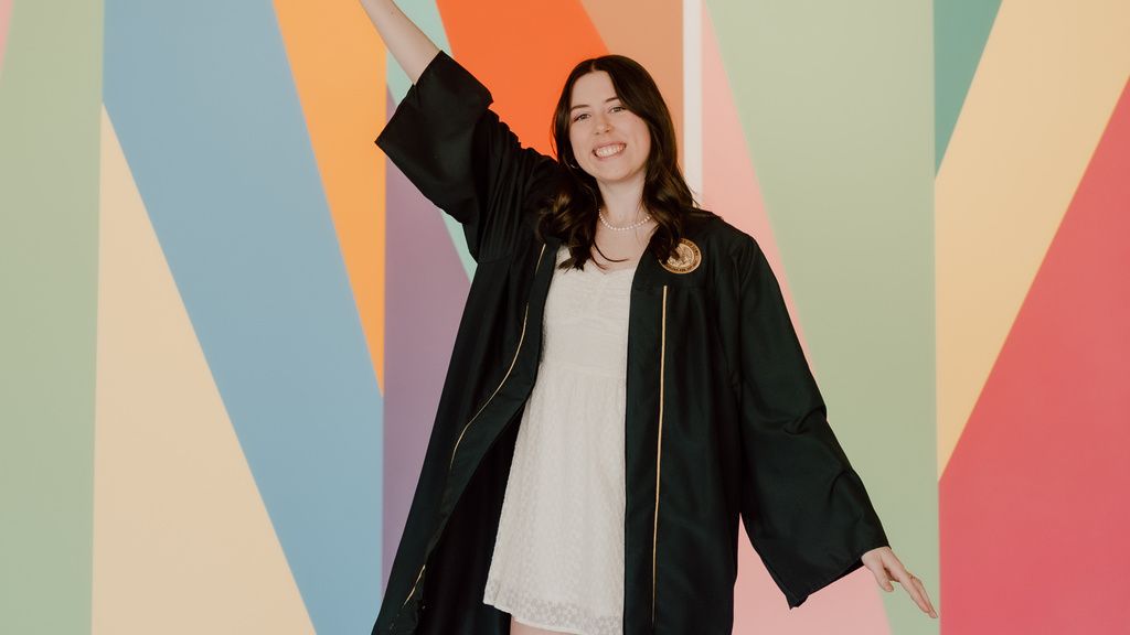 A photo of Anaka Sanders; she is posing in front of the lobby mural at the Stanley, dressed in her graduation robes, tossing her cap in the air.