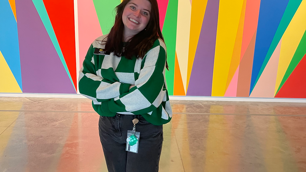 A photo of Alexis Belme: she is standing in front of the mural in the Stanley Museum of Art lobby, smiling, her arms crossed.