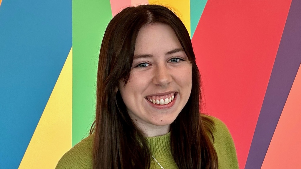 A photo of Anaka Sanders: she is wearing a green knit sweater with a smiley face on the front and standing in front of the colorful mural in the lobby of the Stanley Museum of Art.