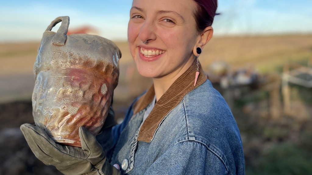 A photo of Eliza Link: she is smiling at the camera, holding a large pot with oven mitts.