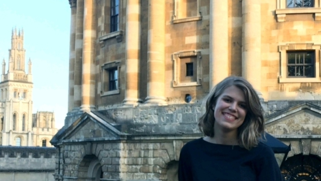 A photo of Maddie Meylor: she is smiling, head tilted, standing in front of an old brick building.