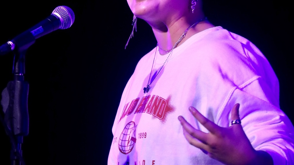 A photo of Samm Yu: bathed in pink light, they are standing in front of a microphone, speaking and gesticulating.