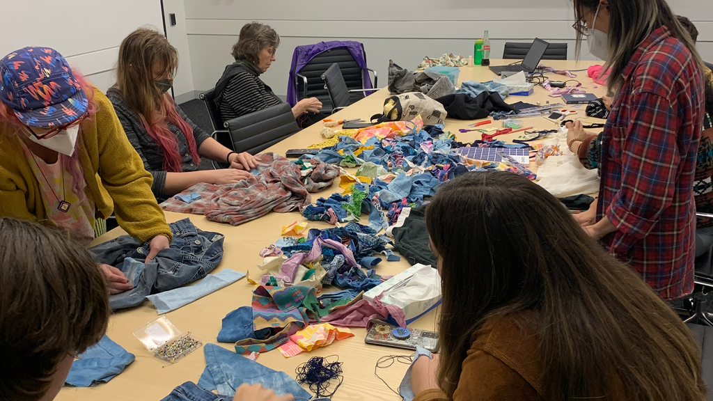 A photo of the class gathered at the table. Fifteen or twenty students sit at a long conference table covered in fabric scraps, books, and garments. At the head of the table is a screen that is displaying a close up of varying sashiko stitches.