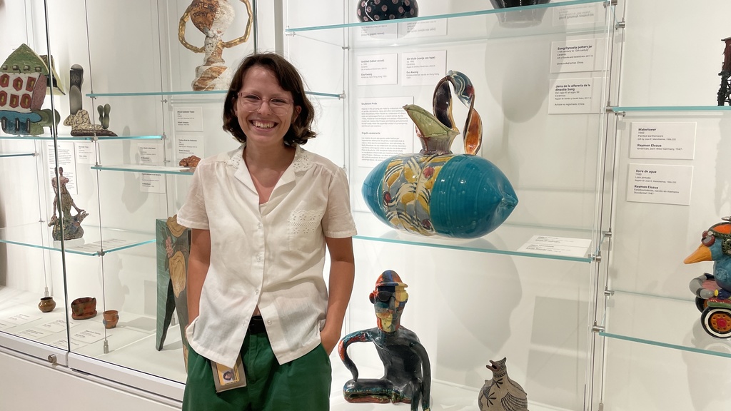 a photo of Melanie, smiling and posing in front of a glass display case of ceramics.