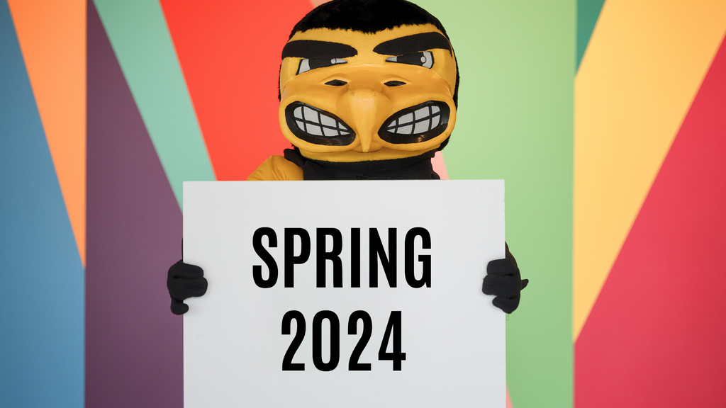 A photo of Herky standing in front of the Stanley's lobby mural, holding a sign that says "Spring 2024"
