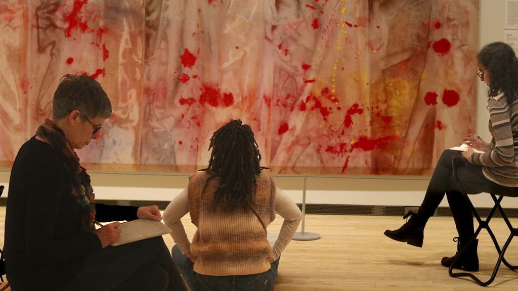 An Image featuring three people sitting in front of Sam Gilliam's "Red April," with notebooks.