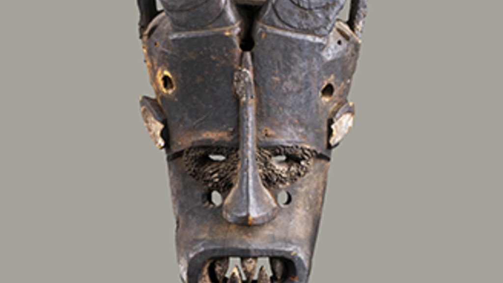 A dark, wooden mask with two large, curved horns that extend upwards and slightly outwards. The face includes a broad nose, large circular eyes, and an open mouth with five large, pointed teeth.
