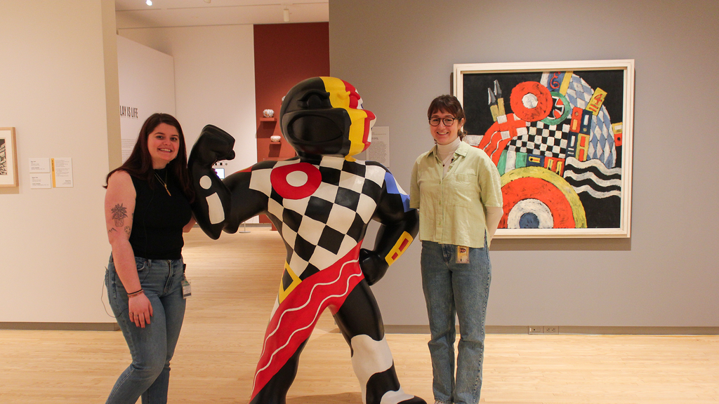 Two students stand in the galleries at the Stanley Museum of Art, posing next to a 6-foot-tall fiberglass Herky statue, that is painted to match a painting in the background of the photo--Marsden Hartley's "E."