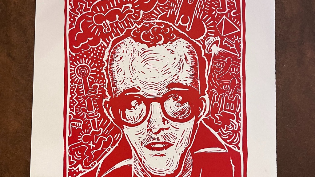 A photo of a linoleum block print; printed in red ink, the image is of Keith Haring, a young man wearing glasses, with vibrant, childlike imagery, reminsicent of his artwork, swirling around his head. 