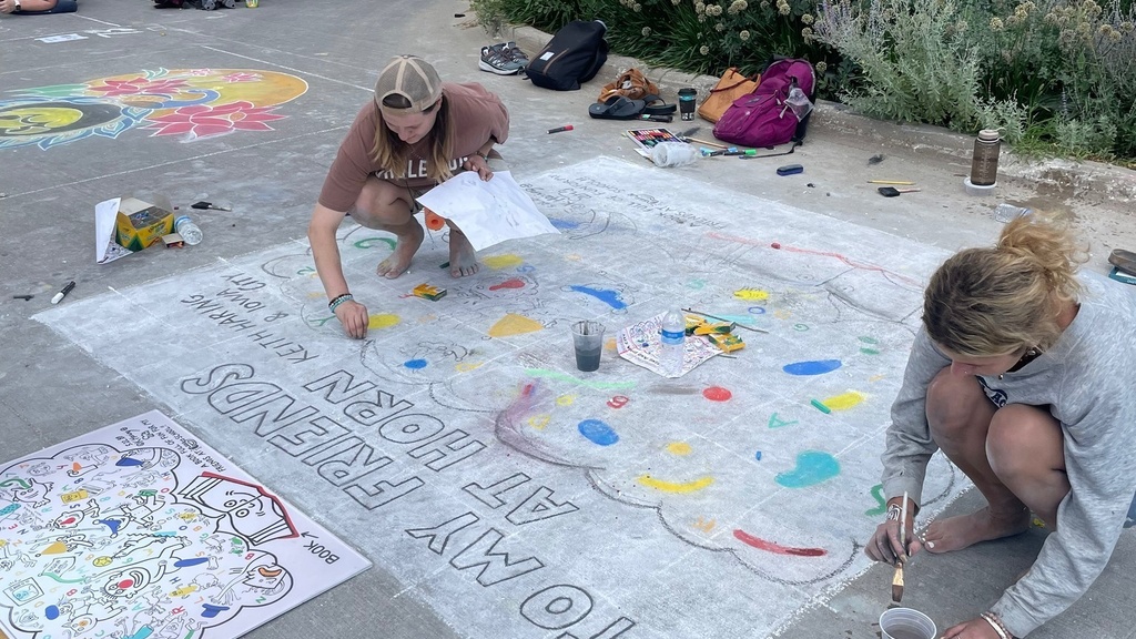 Student employees Sarena Gibson and Mari Johnson crouch on the street in downtown Iowa City, where they are working on a large chalk drawing as part of the city's Rock the Chalk summer festival.