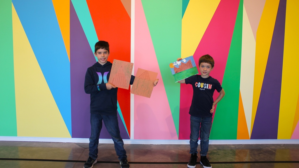 Two boys posing in front of a colorful wall, holding up drawings
