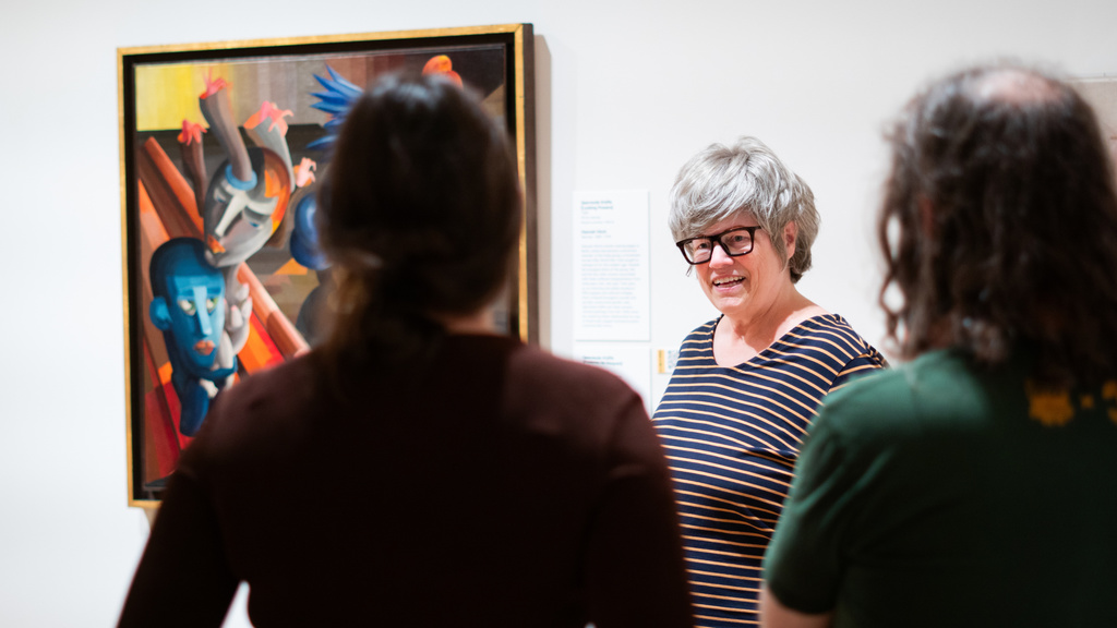 A person with glasses standing in front of a painting, speaking to a group