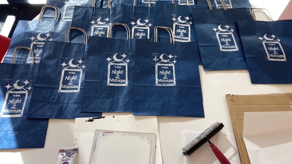 A photo of the entire setup: approximately one dozen printed blue paper bags are lined up along the top of a table, waiting to dry, set above a bottle of white ink, a plate with white ink on it, the ink covered linoleum block, an inked up brayer, and a clean brayer that has been used to apply pressure to the back of the plate to help transfer the ink.