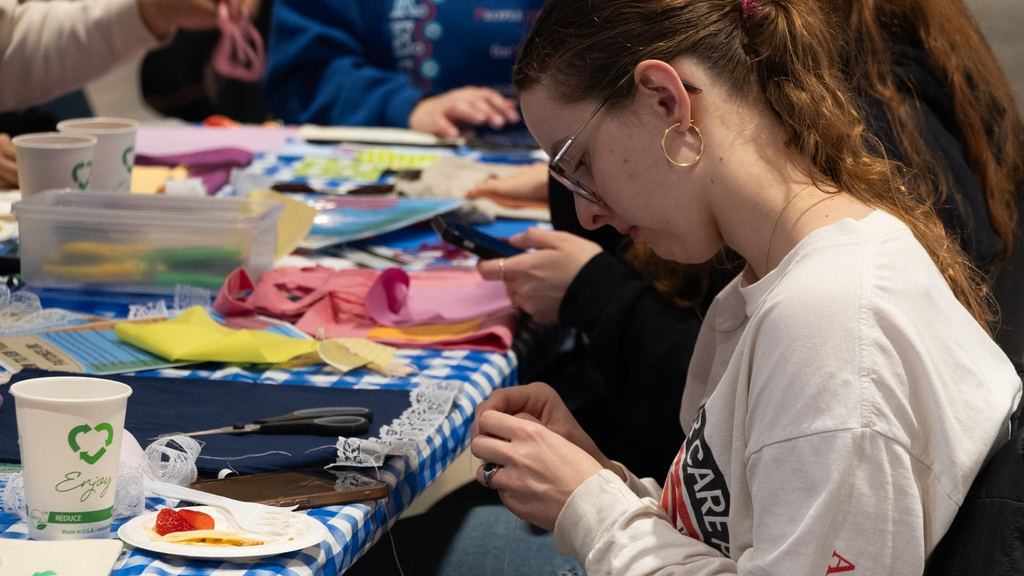A photo of a student, in profile, working on their sewing project; fabrics, sewing supplies, and other materials are scattered in front of them on a table covered with a blue gingham tablecloth.